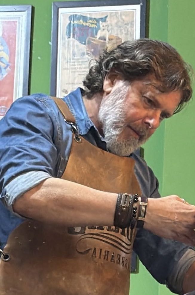 Marcelo, de avental de couro, cortando cabelo diante da parede de quadros