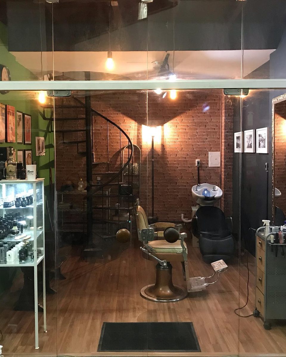 A loja vista pela vitrine: cadeira de barbeiro antiga, parede de tijolo, escada caracol e expositores de vidro